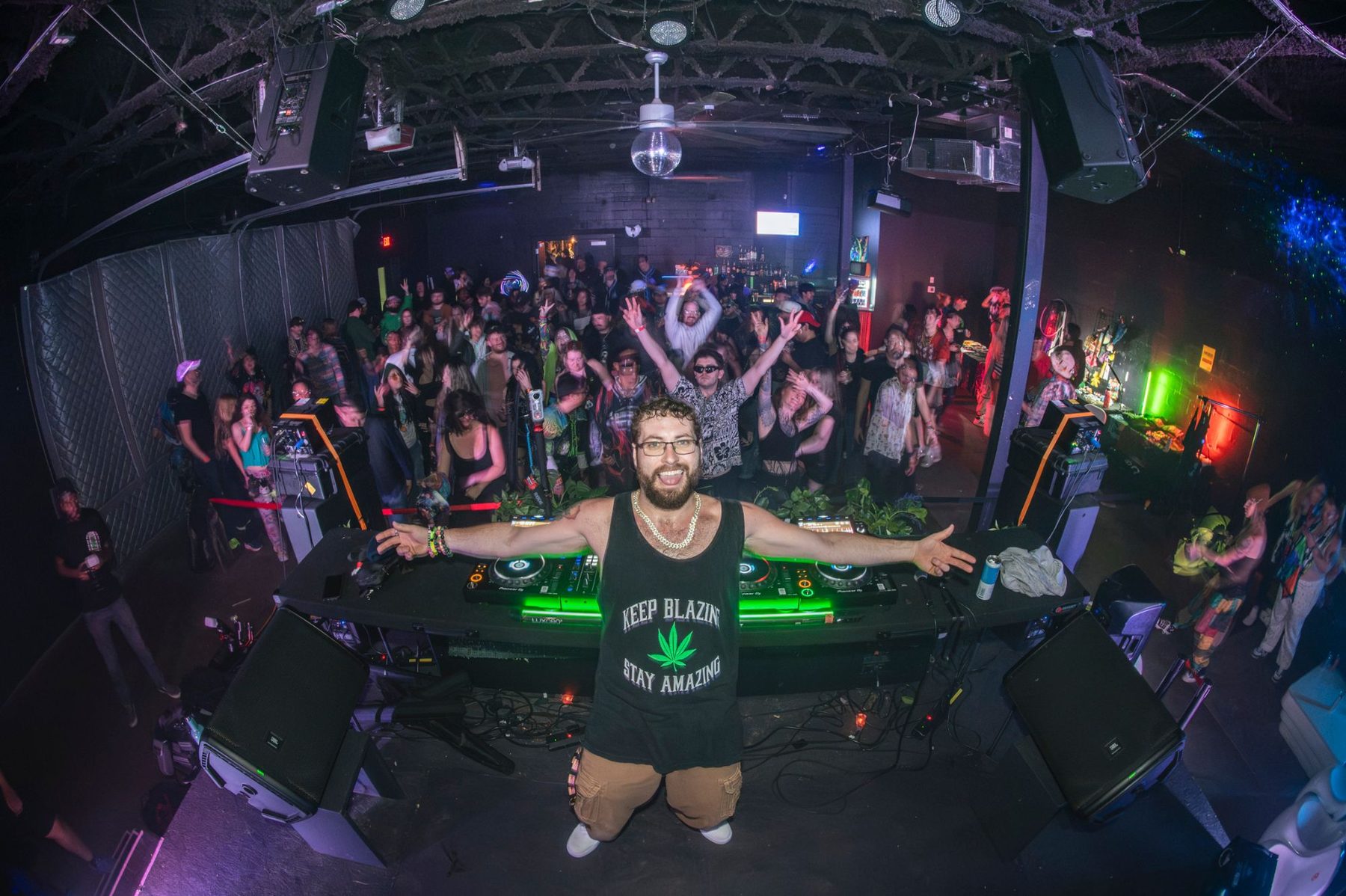 A DJ with arms spread wide behind the booth, a packed crowd dancing in front